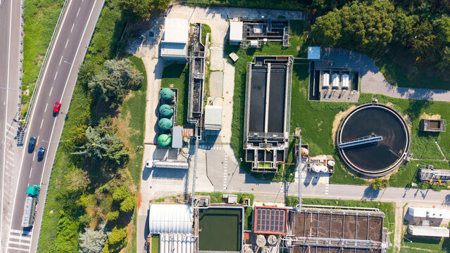 Aerial view of a wastewater treatment plant with circular sedimentation tanks, rectangular aeration basins, and green storage silos, next to a highway in Rancate, Lombardia, Italy.