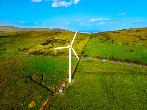 Aerial view of a wind turbine in a green hilly landscape with a road and car under a bright blue sky in Lisburn, Northern Ireland, United Kingdom.