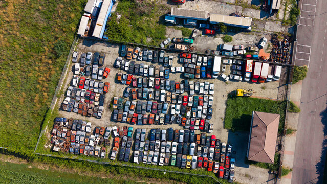 Aerial view of a large salvage yard filled with rows of old cars, scrap metal, and industrial containers in San Giuliano Milanese, Lombardy, Italy.