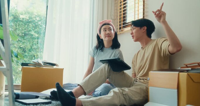 Young Asian couple plans move at home while browsing furniture online on tablet beside packed boxes. Relocation planning, new home setup, online shopping, household design and unpacking concept.