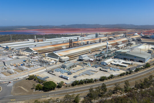 Aerial view of the Boyne Smelter industrial facility with large metal buildings and a red tailings pond in the background in Boyne Island, Queensland, Australia.