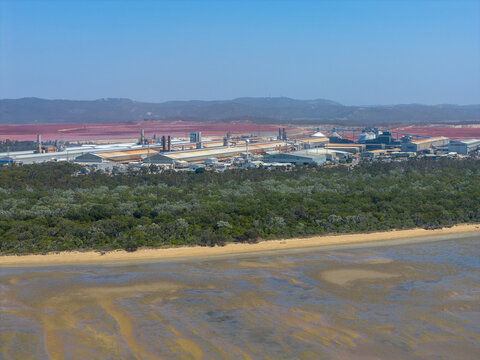 Aerial view of the Boyne Smelter industrial facility situated near the coastline with a red tailings pond, surrounding forest, and sandy beach in Boyne Island, Queensland, Australia.