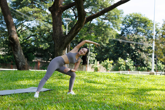 Young Asian woman practicing yoga and stretching outdoors in a green park, performing a side bend pose with a joyful expression, embracing nature and healthy living
