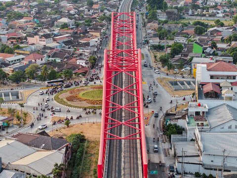 Aerial view of the iconic red steel Jembatan Kereta Api Joglo railway bridge spanning over a busy roundabout with traffic and urban residential surroundings in Surakarta, Jawa Tengah, Indonesia.