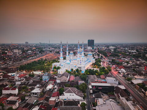 Aerial view of Sheikh Zayed Grand Mosque with white walls and blue domes surrounded by urban rooftops and city streets at dusk in Surakarta, Central Java, Indonesia.