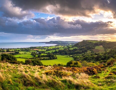 Rolling green hills and fields lead to a serene coastline under a dramatic cloudy sky.