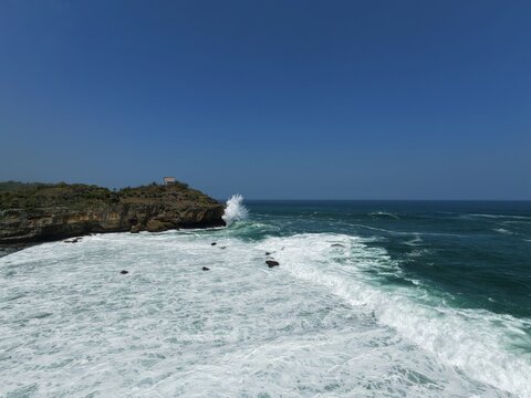 Aerial view of Kasap Beach with powerful waves crashing against rocky cliffs and deep blue ocean waters in Pringkuku, East Java, Indonesia.