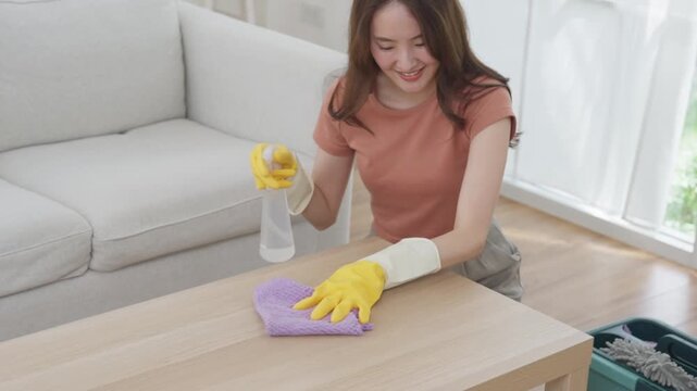 Woman cleaning wooden table with spray bottle, rubber glove, microfiber cloth bright living room home, wiping furniture surface during household chore, smiling indoors, Asian woman cleaning home