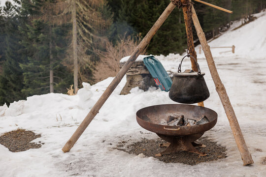 treppiede in legno, braciere rotondo in metallo e un calderone o pentola in ghisa sospeso con una catena sopra il fuoco,  su un terreno innevato, all'aperto, in un ambiente di montagna, in inverno