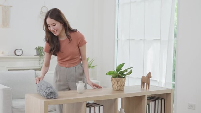 Woman cleaning wooden table with feather duster in bright modern home, houseplant decor, warm smile and calm domestic routine in stylish living room interior