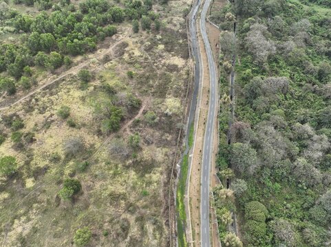 Aerial view of a road winding through a rural landscape with lush green trees and dry vegetation in Cilacap Regency, Central Java, Indonesia.