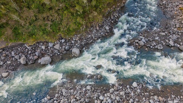 Aerial view of the Blue Rata Reserve, featuring a rushing river with white water rapids flowing over grey stones, flanked by dense green forest in Okato, Taranaki Region, New Zealand.