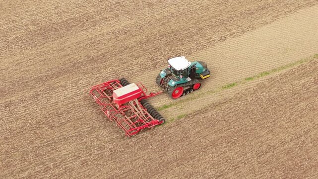 Tracked farm tractor pulling wide seed drill across cultivated soil. Crop sowing machinery working along field rows after harvest. Mechanized agriculture with heavy equipment for commercial grain