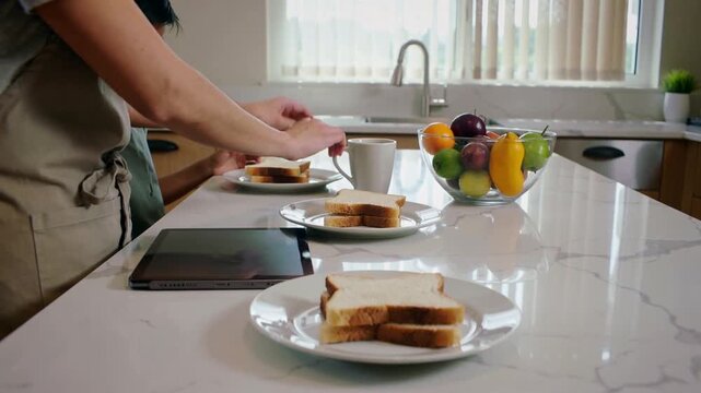Placing, aproned mom urging kid arranging plates on marble island for breakfast with mug
