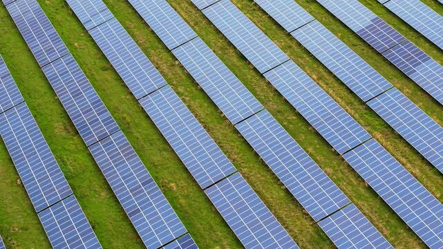 Parallel rows of blue solar panels across green grass from overhead view. Photovoltaic array with geometric pattern in rural field. Utility scale renewable energy installation from aerial perspective