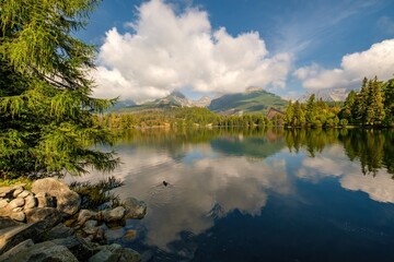 Obraz premium Beautiful scenic view of Strbske pleso lake in High Tatras National Park, Slovakia. Pristine mountain landscape with reflection in water, green forest, rocks, blue sky and dramatic clouds.
