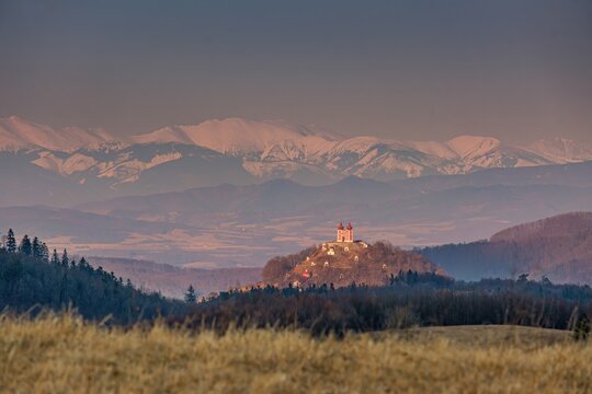 Beautiful scenic view of unesco calvary church on hill in Banska Stiavnica, Slovakia. Historic baroque chapel architecture with distant snow covered tatra mountain peaks during sunset golden hour.