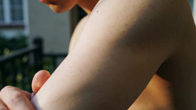 Bringing right hand to left shoulder, shirtless man rubbing down arm on balcony, checking soreness
