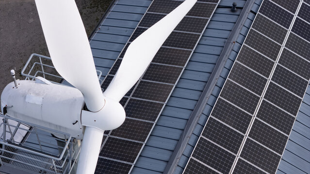 Aerial view of a white wind turbine generator installed next to rows of solar panels on a metal roof in Gdansk, Poland.
