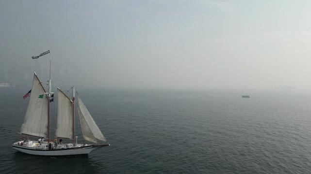 Aerial drone view of two masted schooner sailing across misty Puget Sound, with calm dark water, distant vessel and foggy coastal landscape surrounding boat