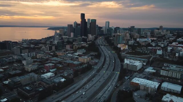 Aerial drone view looking along Interstate 5 toward Seattle downtown with Columbia Center and high rise buildings visible. Elliott Bay and sunset light extend across the background.
