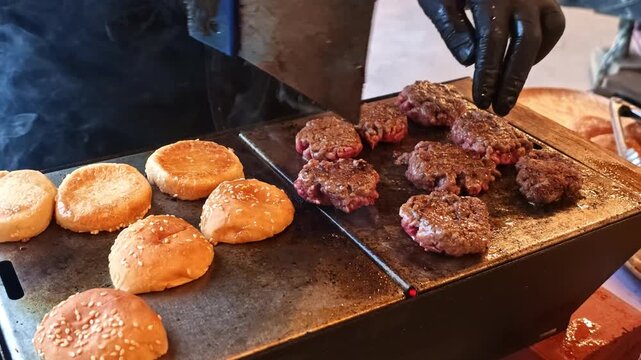 Close up shows gloved hands preparing mini burgers on a hot metal griddle