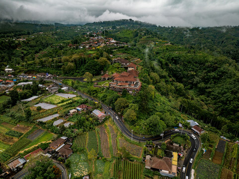 Aerial view of the abandoned Hotel Pondok Indah Bedugul, also known as the Ghost Palace, set amidst lush green hills, terraced fields, and a winding road in Kecamatan Baturiti, Bali, Indonesia.