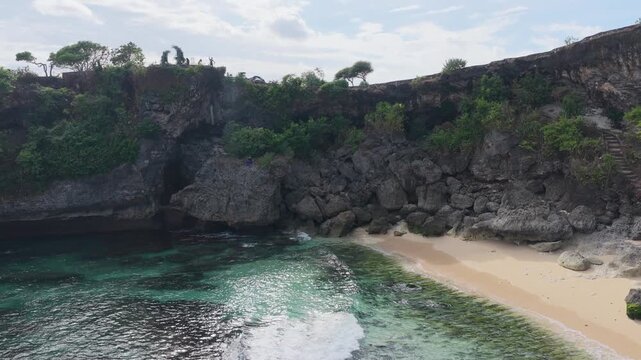 Drone shot moving backward over clear water and a sandy beach, revealing rugged cliffs and the surrounding shoreline at Balangan Beach, Bali, Indonesia