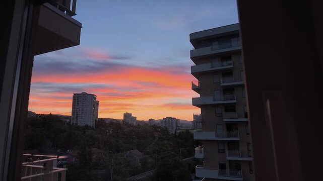vivid sunset viewed from a high-rise apartment through an open window and sheer curtains. The camera slowly reveals a colorful sky with glowing orange and purple clouds above a residential cityscape