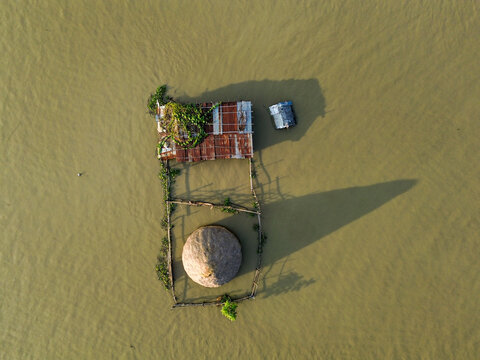 Aerial view of a small hut with a thatched roof and corrugated metal roof partially submerged in floodwaters in Rajshahi District, Rajshahi Division, Bangladesh.