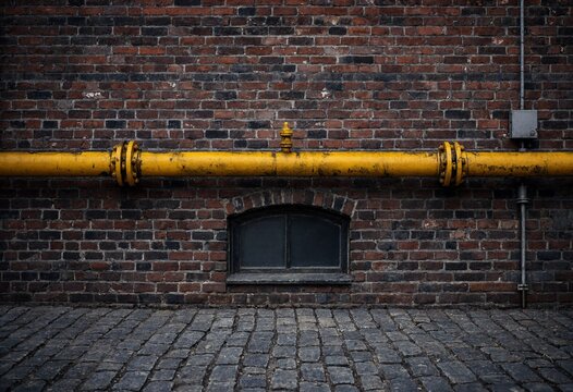 Industrial yellow pipeline running across an old dark brick wall with a small basement window above cobblestone ground