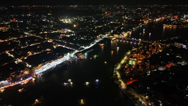 Aerial perspective of Hoi An, Vietnam, highlighting brightly lit streets, colorful architecture, and shimmering reflections on the water at night