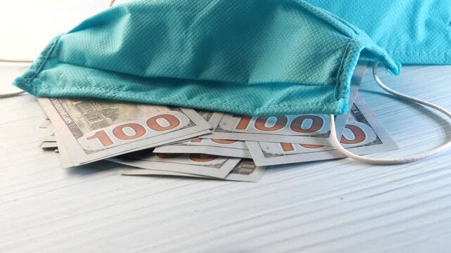 Turquoise protective face mask resting on pile of hundred dollar bills money during pandemic