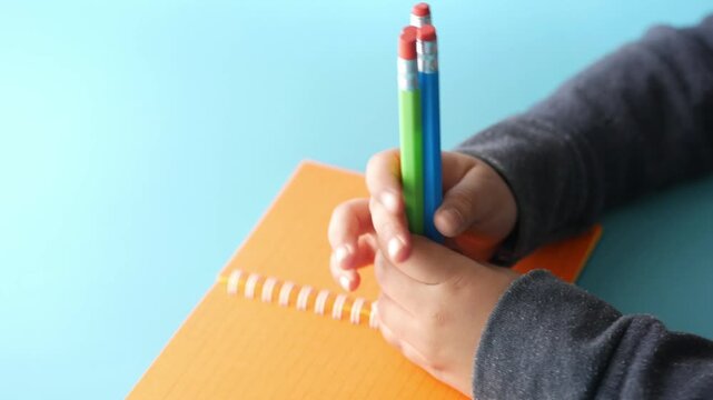 Childs hands holding multiple colored pencils over orange notebook on blue background