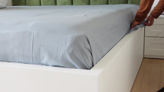 Person tucking in fitted sheet on bed with white frame and green headboard
