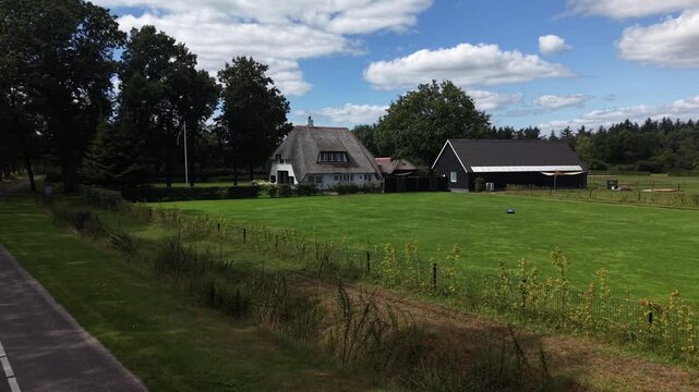 Traditional thatched roof house in Netherlands with white facade, black wooden barn, large green lawn, garden hedges, quiet country road, Dutch countryside under blue sky.Aerial drone approaching shot