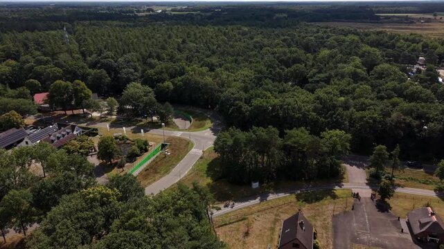 Brick farmhouse with steep gabled roof stands beside thatched barn, gravel courtyard connects outbuildings near curved forest road lined with trees in rural Netherlands. Aerial drone wide shot