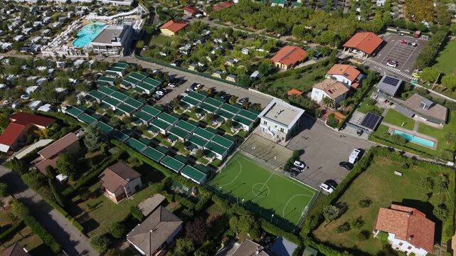 Camping resort at Lake Garda in Italy with rows of green roof bungalows, swimming pool with water slide, soccer field, parking lot, villas with gardens in bright sunlight. Aerial drone top down rising
