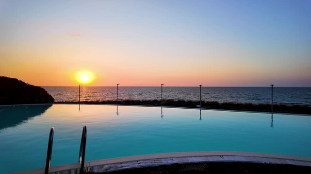 Sunset just above the horizon, over a pool with crystal clear, light blue water, an infinity pool overlooking the calm sea