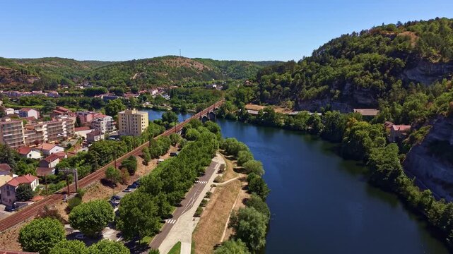 Panorama drone shot near Cahors railway bridge or Le Pont de Chemin de Fer de Cahors spans the Lot River beside a riverside promenade and residential blocks on a sunny summer day, France