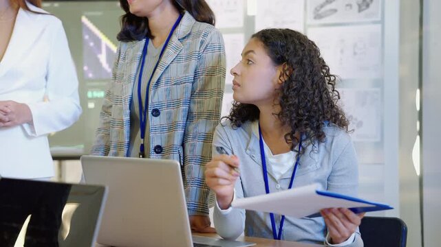 Female engineering professional taking notes during a collaborative technology meeting while working with colleagues, digital devices, and project documents in a modern office.
