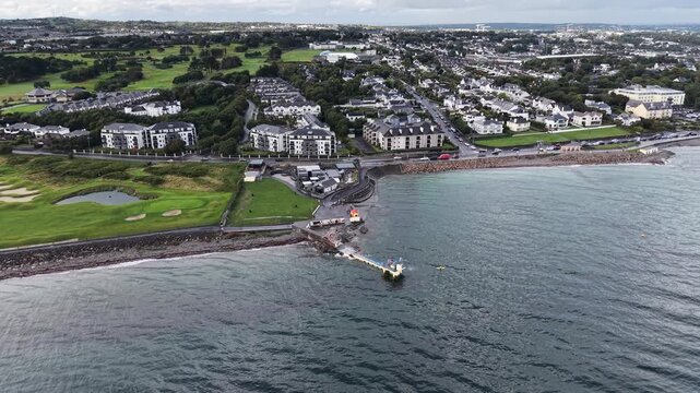 Wide aerial drone view of Salthill in Galway, Ireland, showing the coastline, rocky shore, Galway Bay, coastal road, residential neighborhoods and green golf course. The panoramic perspective captures