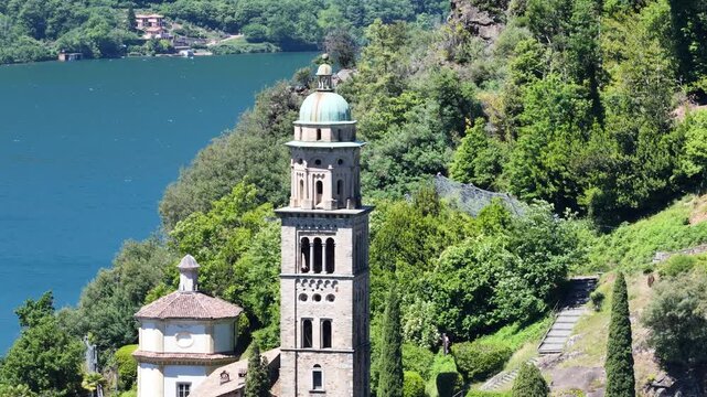 Scenic view of the historic Santa Maria del Sasso church in Morcote, Ticino, Switzerland, with its distinctive bell tower rising through lush hillside vegetation above the blue waters of Lake Lugano.