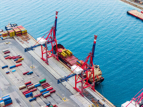 Aerial view of a large container ship docked at the commercial port with red cranes loading cargo containers and stacks of colorful shipping containers on the quay in Lemesos, Lemesos, Cyprus.