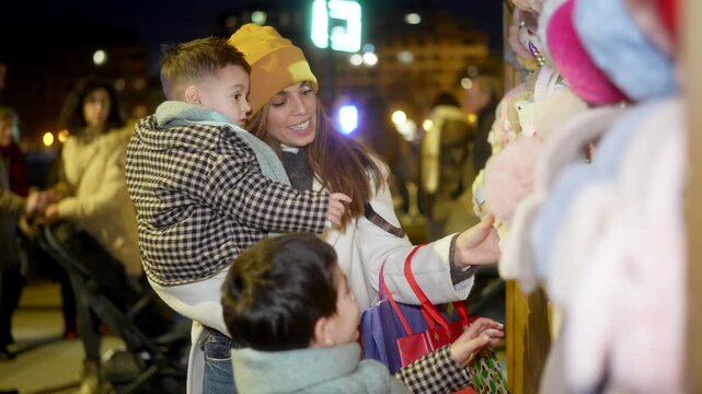 Smiling mother holding her son and shopping with her other child at an outdoor christmas market at night, looking at a stall with toys and holiday gifts creating a festive family memory