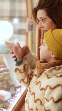 Happy mother and her young son enjoying a magical christmas evening, looking with wonder at a beautifully decorated store window display with festive lights and holiday gifts