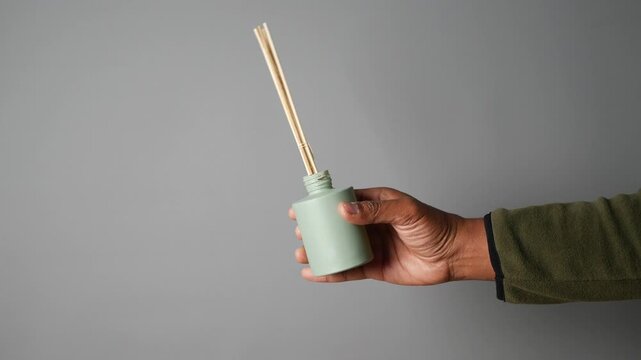 Close up of person holding aroma oil bottle with essential oil diffuser sticks on gray background in a studio shot
