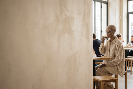 Alien sitting at cafe table in modern bright restaurant with large empty wall copy space