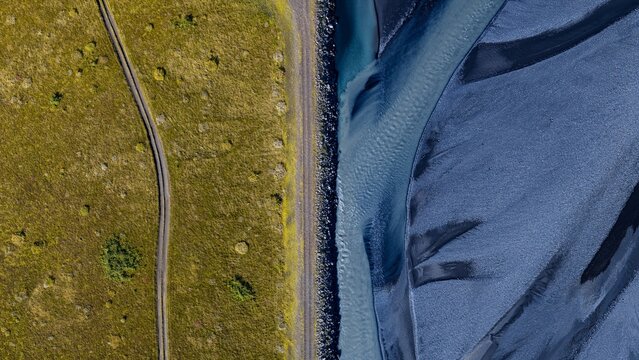 Aerial view of the Markarfljot glacial river featuring a striking contrast between the vibrant green grassy bank with a dirt track and the dark, braided riverbed in Rangarthing eystra, Iceland.