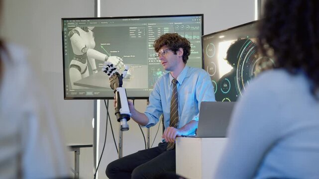 Engineer demonstrating a robotic arm prototype during a technology presentation while colleagues observe, with digital displays showing robotics data and humanoid robot concepts.

